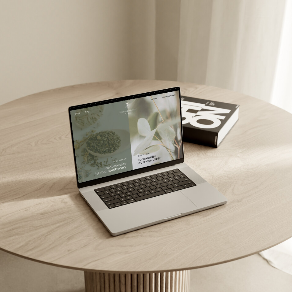 roncys apothecary and clinic website home page mockup on macbook laptop on wooden circular table with black coffee table book and neutral background